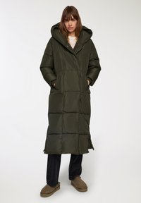 Cappotto lungo e soffice di colore verde con cappuccio, caratterizzato da un design a collo a V, ampie tasche anteriori e una texture trapuntata. Indossato sopra pantaloni scuri e calzature.