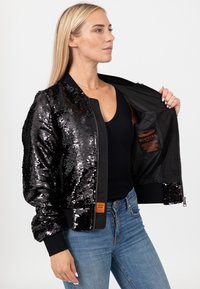 Vrouw houdt een open zwarte bomberjas met pailletten vast waarin een oranje label zichtbaar is, draagt een zwarte top en een blauwe spijkerbroek tegen een witte achtergrond.