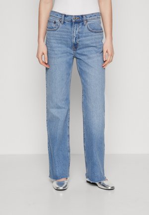 Person wearing light blue straight-leg jeans with raw hems and silver low-heeled shoes, standing against a plain background.