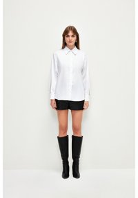 Camicia bianca con colletto classico, maniche lunghe e texture liscia, abbinata a pantaloni corti neri e stivali neri fino al ginocchio.