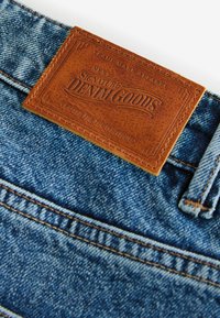 Blugi din denim cu un eticheta din piele maro texturată, cu text în relief, detalii cusute cu fir în contrast și o spălare clasică albastră.