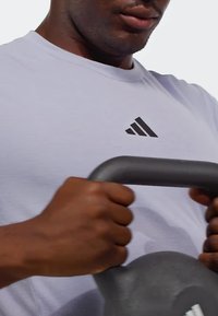 Kettlebell gris sostenido con ambas manos, con el logo negro de Adidas en una camiseta deportiva gris clara. Textura suave, diseño redondeado.