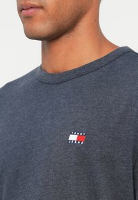 Tumši pelēks kokvilnas T-krekls ar apaļu kakla izgriezumu, kurā kreisajā krūtīs ir neliels izšūts logo zilos, sarkanos un baltos toņos.