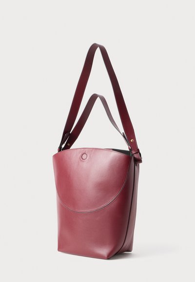 Bolso de cuero burdeos con una parte superior redondeada, correa de hombro ajustable y una textura suave. Presenta un acento circular en la parte frontal.