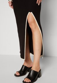 Robe maxi noire côtelée avec une fente latérale bordée de crème. Portée avec des mules noires à talons ouverts dotées d'un talon épais.