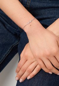 Silverarmband med tre cirkulära klara ädelstensaccenter, länkade av en delikat kedja, som bärs på en hand som vilar på mörk denimtyg.