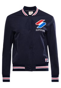 Marinfärgad bomberjacka i slätt tyg. Har röd och vit randig ribbad krage och manschetter, med broderad logotyp på bröstet och sidan.