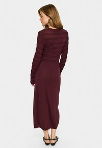 Long maroon dress with a fitted, textured bodice, long sleeves, and a flowing lower portion, featuring back detailing and ankle-length hem.
