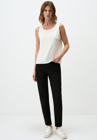 Weißes ärmelloses Oberteil, schwarze enganliegende Hose und weiße Sneakers. Das Oberteil hat einen runden Halsausschnitt; das Outfit ist einfach und modern im Design.