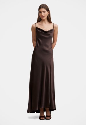 Mujer de pie con un vestido largo de satén marrón oscuro con tirantes finos y tacones abiertos marrones sobre un fondo liso.