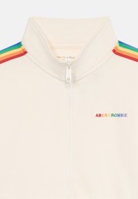 Giacca bianca con zip, caratterizzata da un colletto alto, accenti a strisce colorate in rosso, arancione, giallo, verde e blu sulle spalle, e logo ricamato.