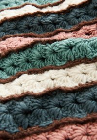Pătură croșetată cu fire groase și texturate în culori alternante: verde teal, crem, roz și accente maro închis, cu dungi ondulate.