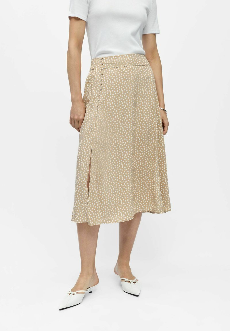 Jupe midi beige en ligne A avec des pois blancs irréguliers, fente latérale, portée avec un top blanc à manches courtes et des chaussures blanches à enfiler à bout pointu.