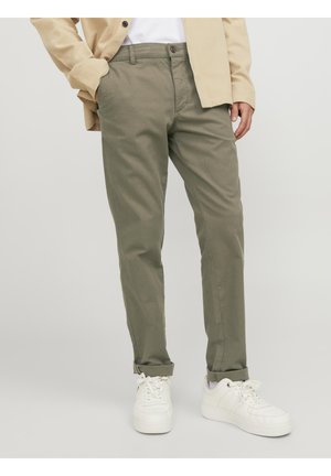 Man draagt opgerolde olijfgroene broek, beige jas, wit shirt en witte sneakers, staand tegen een effen achtergrond.