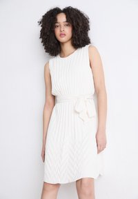 Pleated sleeveless dress in off-white, featuring a belted waist with a bow tie. The texture includes vertical and chevron patterns.