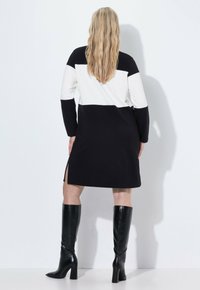 Schwarz-weißer farbblockierender Kleid mit langen Ärmeln, seitlichen Schlitzen und lockerer Passform. Kombiniert mit knielangen schwarzen Stiefeln.