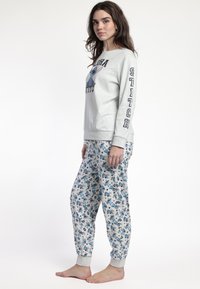 Mujer vestida con sudadera gris y pantalones de pijama a juego con estampado de personaje de dibujos animados azul, de pie descalza contra un fondo blanco.