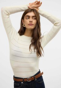 Top a maniche lunghe a coste di colore crema con strisce orizzontali di larghezze variabili; presenta un collo rotondo ed è abbinato a una cintura marrone e jeans scuri.