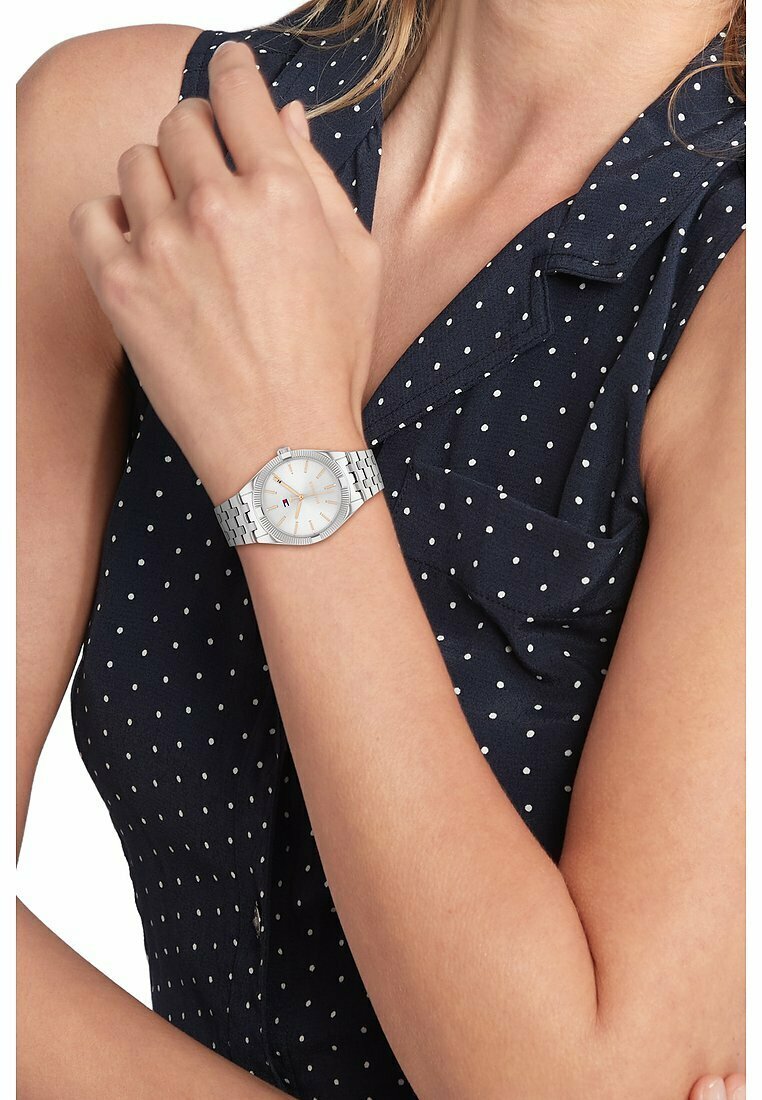 Reloj de pulsera de plata con una caja redonda y texturizada, esfera de color claro y pulsera de eslabones plateados; cuenta con manecillas delgadas y marcadores de hora.
