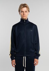 Hombre rubio con pelo corto que lleva una chaqueta deportiva azul marino con cremallera y rayas beige en las mangas, y pantalones a juego con cordones blancos.