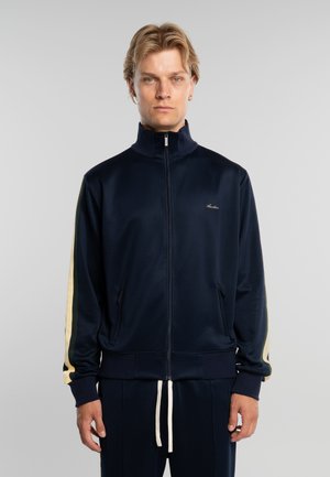 Hombre rubio con pelo corto que lleva una chaqueta deportiva azul marino con cremallera y rayas beige en las mangas, y pantalones a juego con cordones blancos.