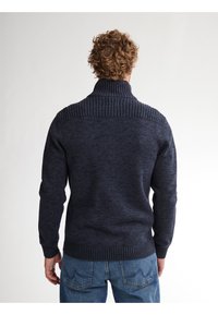 Dunkelblauer Strickpullover mit hohem Rippkragen, gerippten Schultereinsätzen und einer strukturierten Oberfläche. Kombiniert mit hellblauen Jeans.