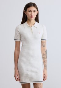 Λευκό πλεκτό φόρεμα πόλο με υφασμάτινο μοτίβο διαμαντιού, κοντά μανίκια με σκούρο περίγραμμα και διακριτικό λογότυπο στο αριστερό μέρος του στήθους.