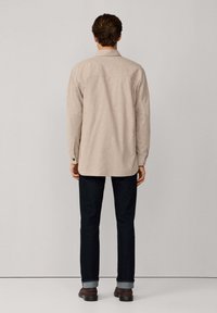 Camisa beige de manga larga con un corte relajado, detalle en la yugo trasero y puños abotonados, combinada con jeans oscuros y zapatos marrones.