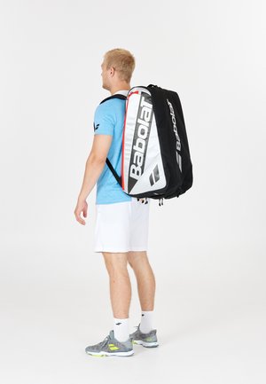 Uomo che indossa una camicia blu, pantaloni corti bianchi e scarpe da ginnastica grigie, con una grande borsa da tennis Babolat nera e bianca sulla schiena, su sfondo bianco.
