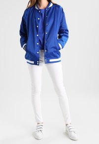 Chaqueta bomber azul con puños y cuello de rayas blancas, cierre de botones, combinada con una camisa azul, pantalones blancos y zapatillas blancas.