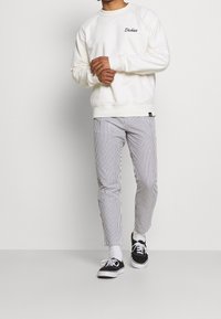 Hvid sweatshirt med ribbet kant og manchetter, med sort broderet tekst; parret med stribede bukser og sorte sneakers.