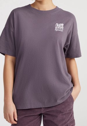 Osoba nosząca luźny fioletowy t-shirt z krótkim rękawem z małym białym graficznym tekstem na lewej piersi oraz pasujące fioletowe spodnie sztruksowe.