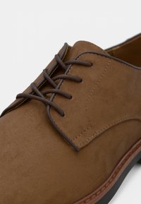 Chaussure à lacets en daim marron avec une texture lisse, des lacets marron contrastants et une semelle noire durable. Détail de couture le long du bord.
