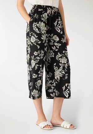 Donna che indossa pantaloni neri a gamba larga e corti con stampa floreale bianca e sandali bianchi slip-on, in piedi con le mani nelle tasche.