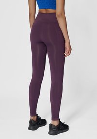 Hoch taillierte lila Leggings aus dehnbarem Stoff, mit glatter Textur und figurbetontem Design; kombiniert mit schwarzen Sportschuhen.