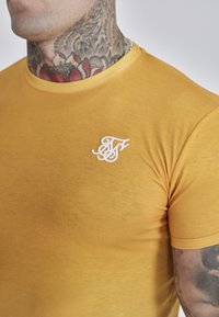 Sennepsgul bomulds T-shirt med rund halsudskæring, korte ærmer og et hvidt broderet logo på venstre brystområde.