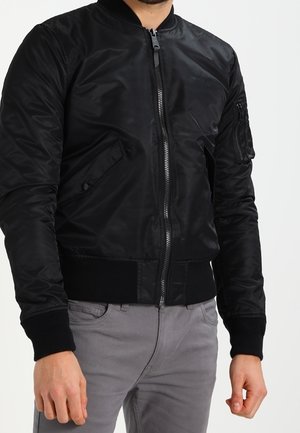 Homme portant une veste bomber noire avec fermeture éclair à l'avant et poches, associée à un pantalon gris, sur un fond clair uni.