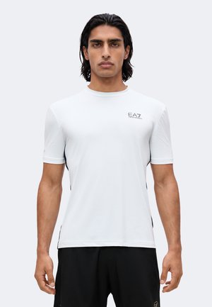 Jeune homme aux cheveux foncés portant un t-shirt de sport blanc à manches courtes EA7 Emporio Armani et un short noir, debout devant un fond uni.