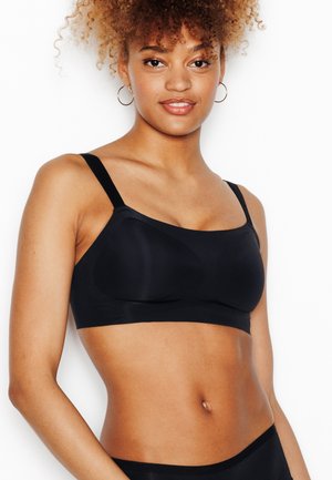 Femme aux cheveux bouclés portant un soutien-gorge noir sans coutures avec bretelles réglables et des boucles d'oreilles en or, debout devant un fond blanc.