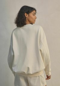 Sweatshirt couleur crème avec une coupe décontractée, épaules tombantes, manches et ourlet côtelés. Porté par-dessus un short à rayures claires. Tissu doux et texturé.
