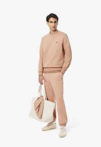 Felpa rosa chiaro e pantaloni joggers abbinati in morbido tessuto. Presenta un logo verde sul petto e un tessuto decorato drappeggiato su una borsa beige.