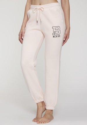 Woman wearing light pink jogger pants with elastic cuffs and black letter "B MCR'89" logo on right thigh, standing barefoot.