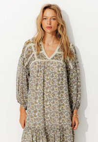 Robe à imprimé floral en tissu léger, avec un décolleté en V, des manches bouffantes longues et un ourlet à volants. Les couleurs incluent crème, rose et jaune.