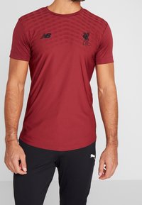 Röd atletisk t-shirt med New Balance-logotyp på bröstet, Liverpool FC-emblem och texturerat mönster på den övre framsidan. Korta ärmar.