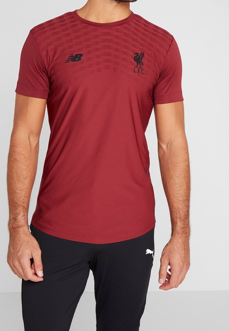 Röd atletisk t-shirt med New Balance-logotyp på bröstet, Liverpool FC-emblem och texturerat mönster på den övre framsidan. Korta ärmar.