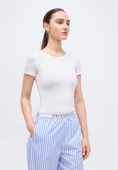Γυναίκα με μαλλιά μαζεμένα πίσω φοράει λευκό εφαρμοστό κοντομάνικο τοπ και παντελόνι με κάθετες μπλε και άσπρες ρίγες, με ζώνη Polo Ralph Lauren.