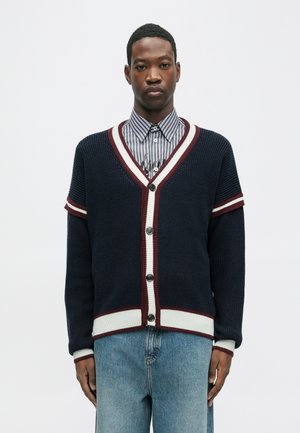 Navy knitted cardigan with maroon and white striped accents, V-neck, and five buttons, paired with a collared striped shirt and denim jeans.