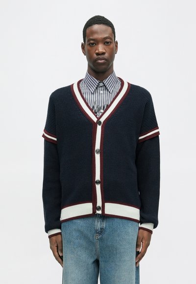 Cardigan en tricot bleu marine avec des accents rayés bordeaux et blancs, en V, et cinq boutons, associé à une chemise rayée à col et un jean en denim.