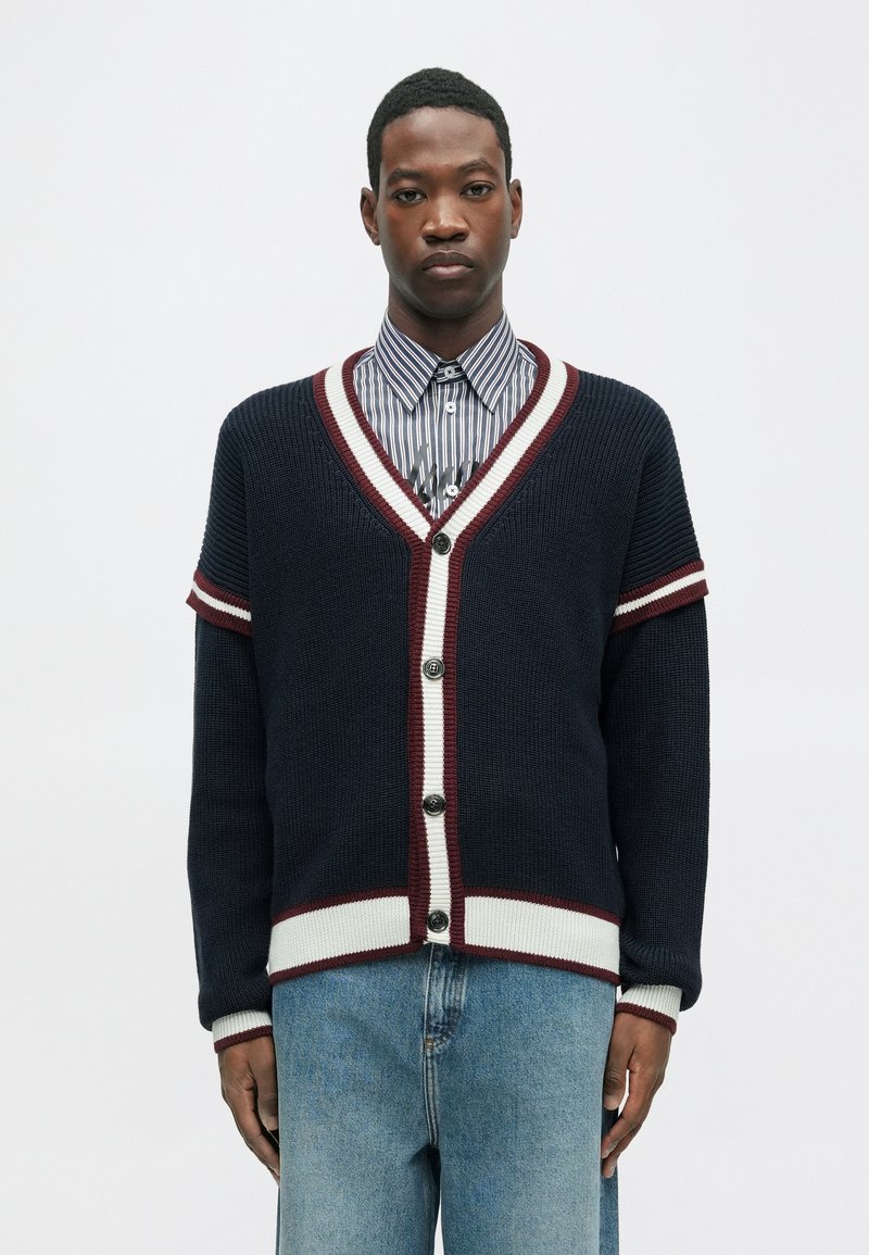 Cardigã de malha azul marinho com detalhes em listras bordô e brancas, decote em V e cinco botões, combinado com uma camisa listrada de colarinho e jeans de ganga.