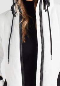 Veste matelassée blanche avec des accents noirs, dotée d'une fermeture éclair et de cordons ajustables sur la capuche, réalisée en tissu lisse.